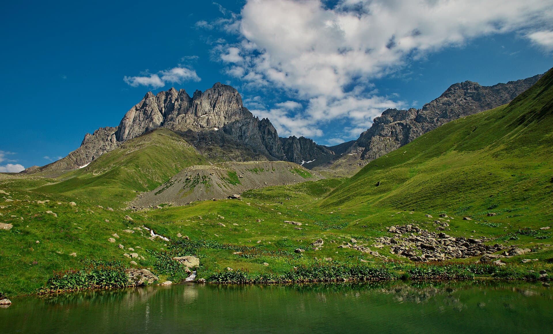 Jagged peaks of the Chaukhi massif above green meadows and a small alpine lake near Juta